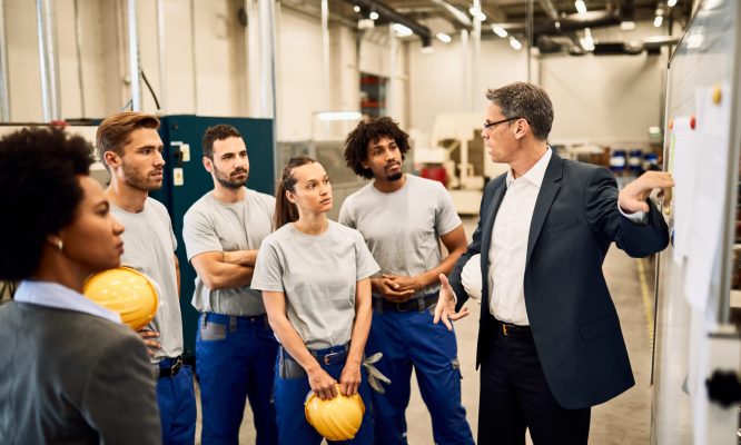 Mid adult businessman giving presentation to group of industrial workers in a factory.