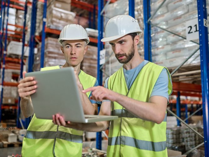 Important point. Two male colleagues in white helmet and bright vest looking attentively at laptop deciding work question standing at industrial warehouse